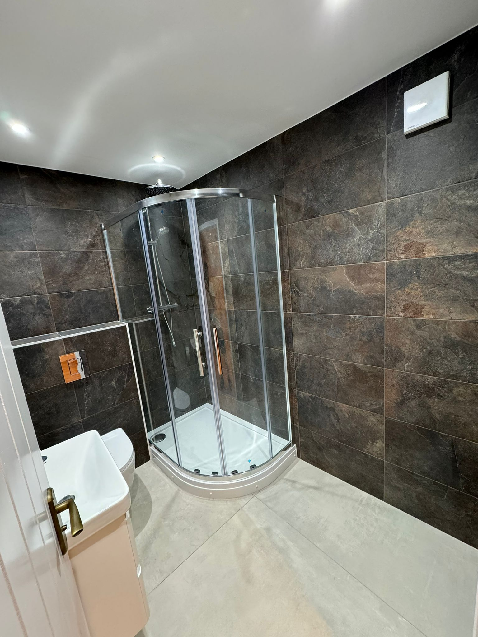 Compact bathroom renovation featuring curved corner shower enclosure, dark slate wall tiles, light grey floor tiles, and vanity sink