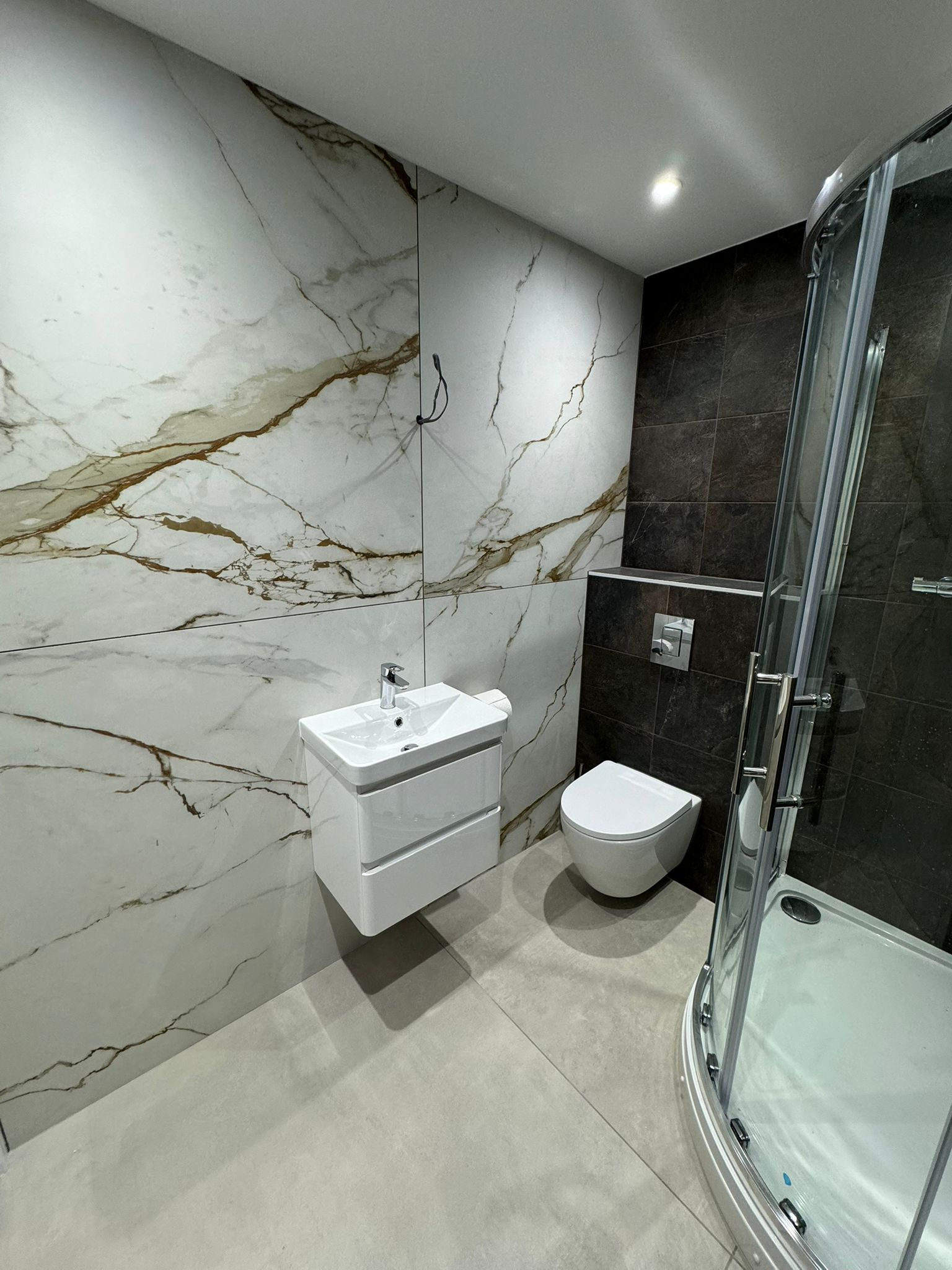 Luxury bathroom renovation featuring white calacatta gold marble wall tiles, dark accent tiles, floating vanity sink, wall-mounted toilet, and corner glass shower