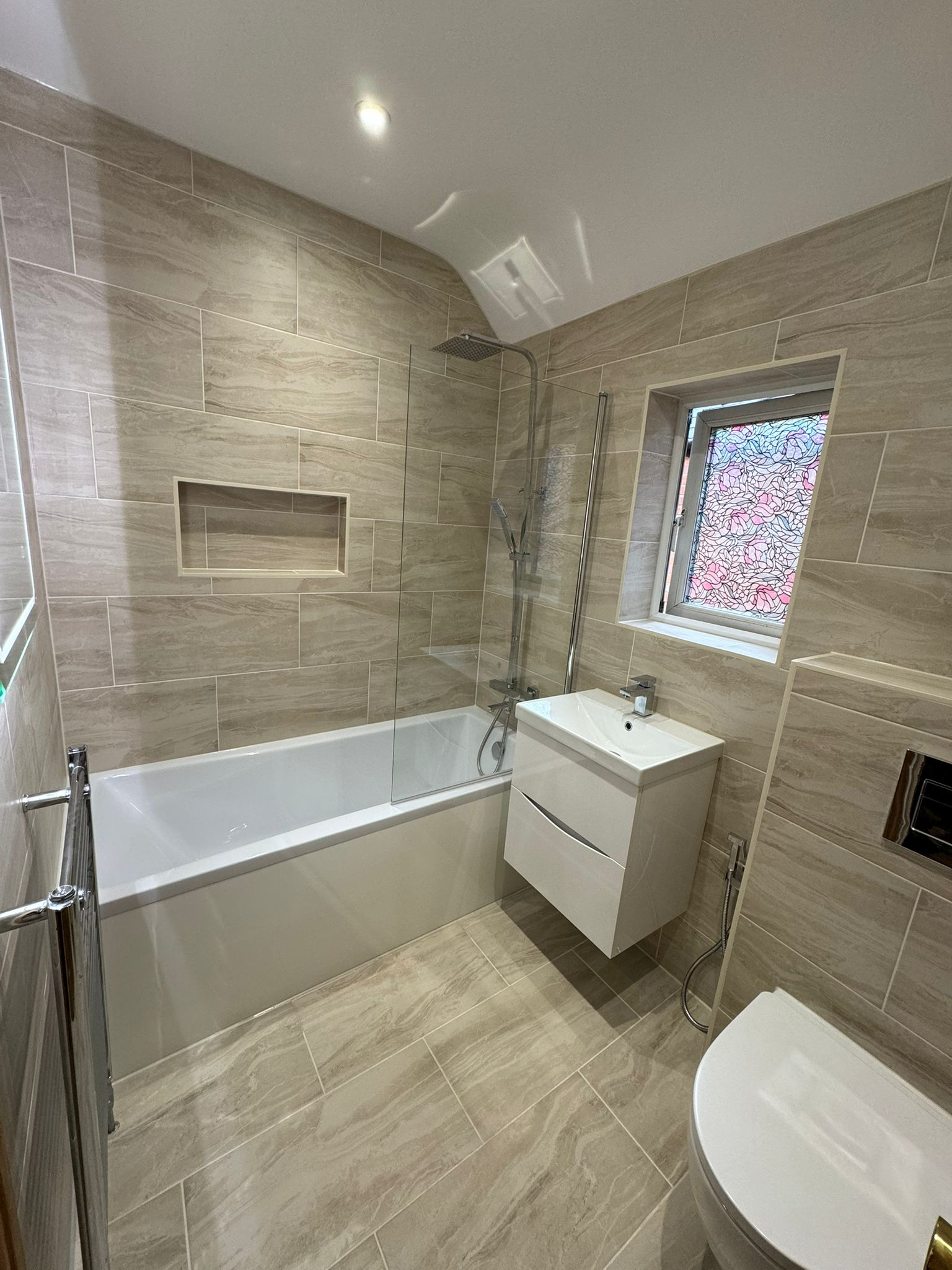 Modern bathroom fitting featuring beige porcelain tiles, bathtub shower combination, glass screen, and floating white vanity