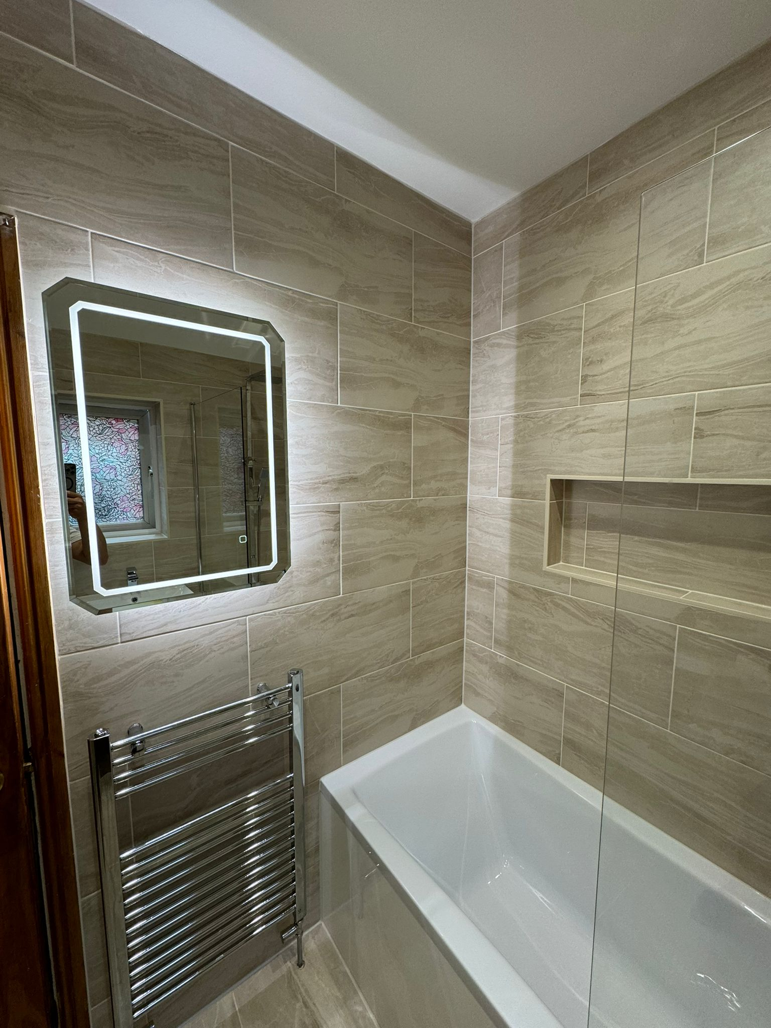 Modern bathroom renovation featuring beige ceramic wall tiles, LED backlit mirror, chrome heated towel rail, and white bathtub with glass screen